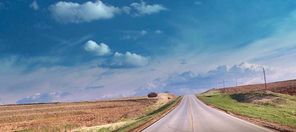 A long paved road through the countryside of Nebraska cuts through a field of dried corn ready for harvest.